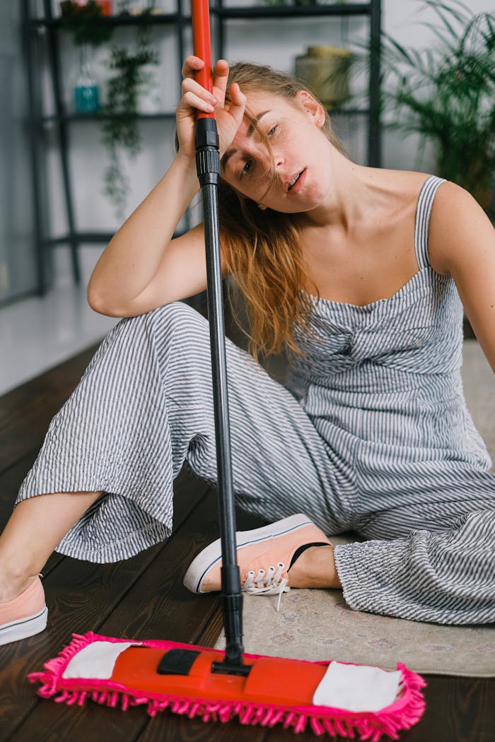 Exhausted young female leaning on handle of mop while looking away and sitting on carpet after housework