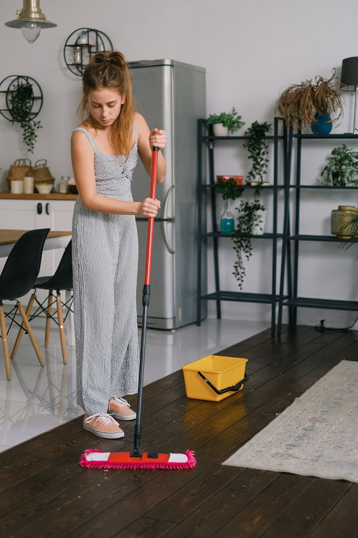 Young female in jumpsuit cleaning parquet with mop against rug and bucket in house room