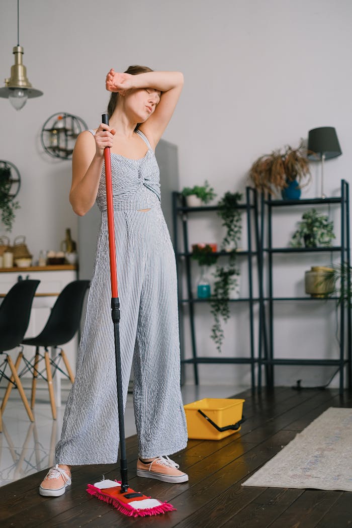 A young woman in a modern home taking a break from cleaning, touching her forehead, illustrating fatigue.